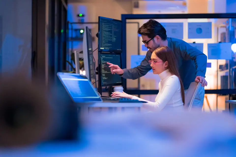 Asian programmers collaborating on software development at night, focused on writing code across multiple monitors in a tech office environment…
