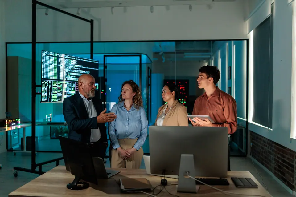 Four IT professionals discuss computer systems and monitoring data together in a modern network operations center, with one team member holding a tablet