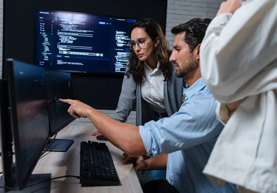 Programmer team discussing cybersecurity workflow and software review at monitor, focused on AI integration, coding review, and problem solving in…