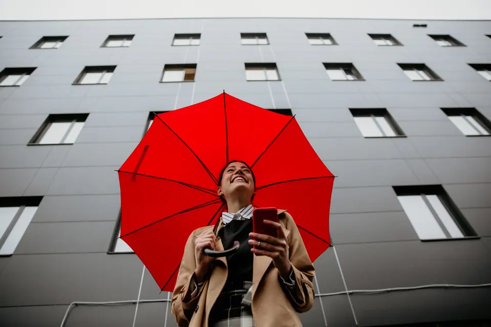 Rear View of a smiling pleased business dressed female with an opened red umbrella and a smartphone standing against the modern gray building