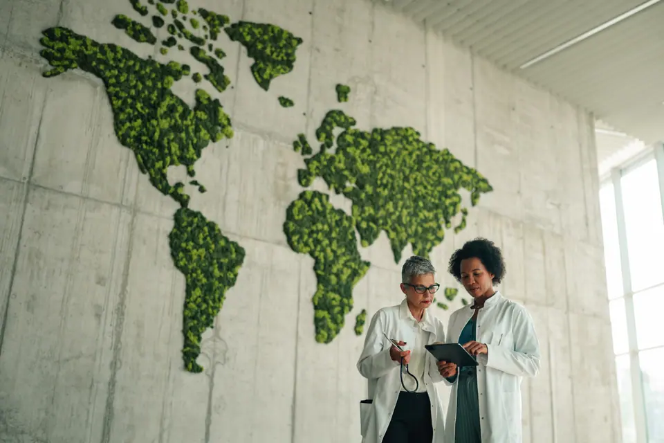 Two female doctors discussing information on a digital tablet in front of a green world map, symbolizing global health and sustainability
