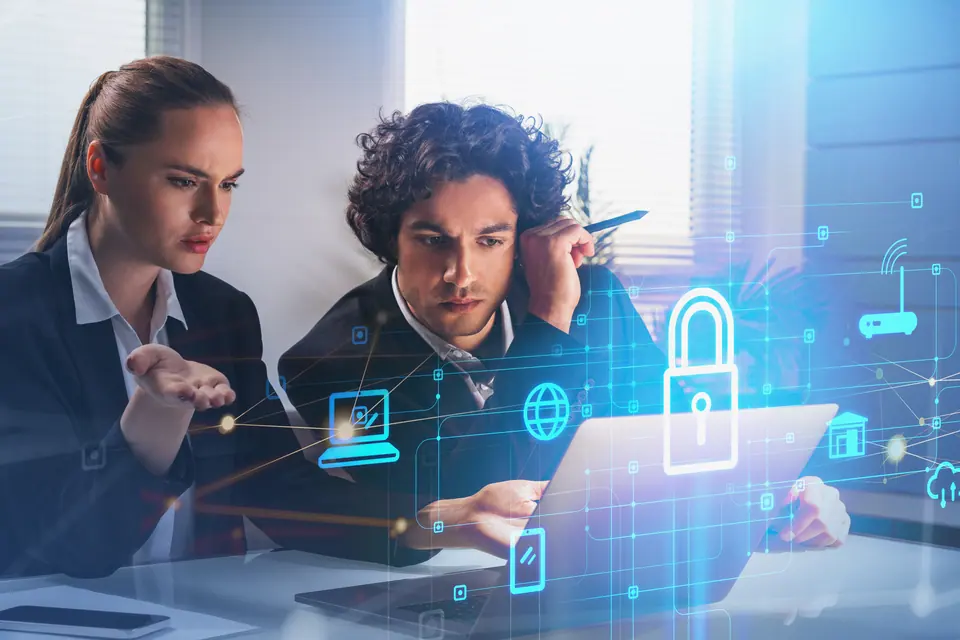 Two thoughtful business people with laptop, desktop at office interior, blue glowing information protection icons