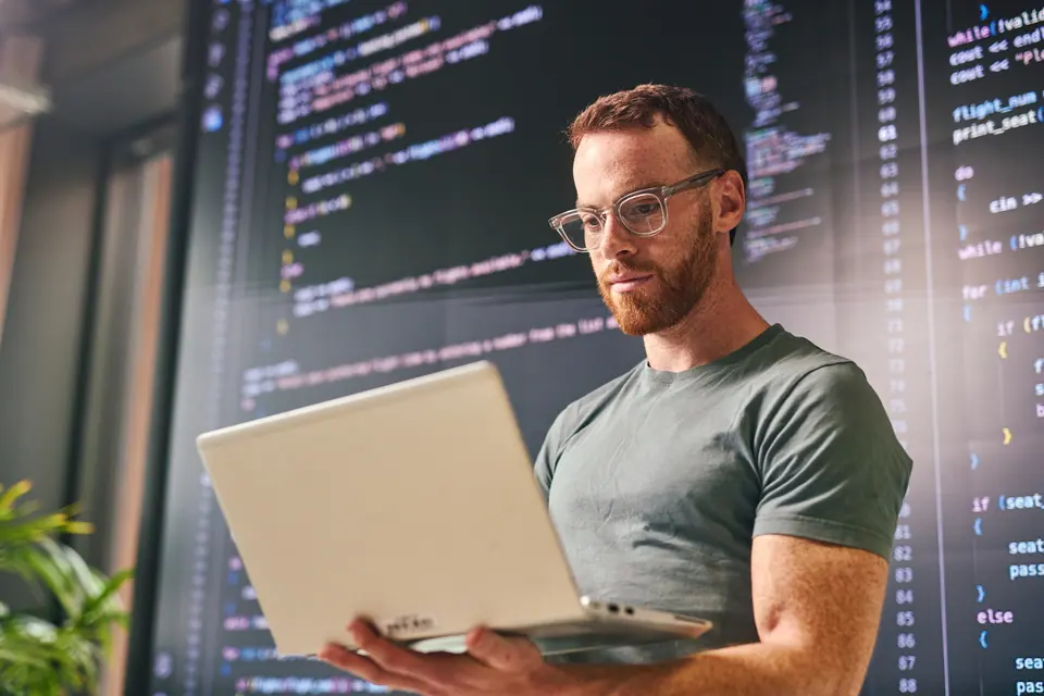 Young adult male software developer concentrating on his laptop, with lines of code displayed on a large screen in the background, in a contemporary…