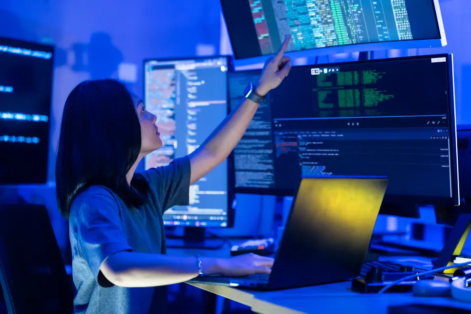 Young Asian woman points toward programming data on upper computer monitor while working at laptop workstation, reviewing source code for cybersecurity…