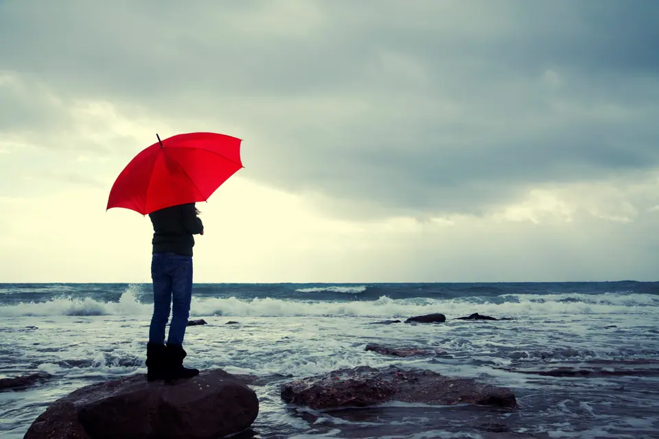 Young girl with red umbrella standing on a stone at the beach and looking away